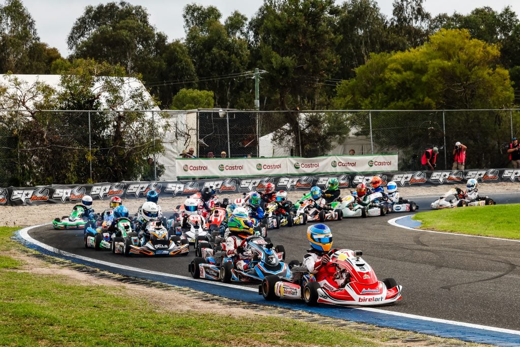 A line of go-karts with drivers in colorful helmets and racing suits is navigating a turn on an asphalt race track.