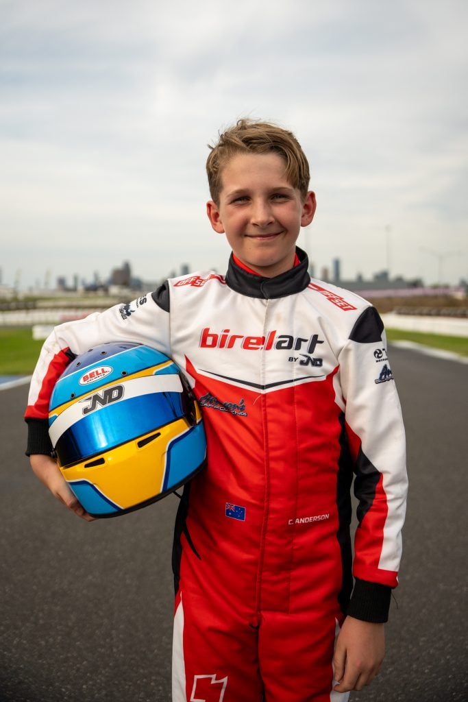 A young boy in a white and red racing suit holds a blue, yellow, and white helmet under his arm.